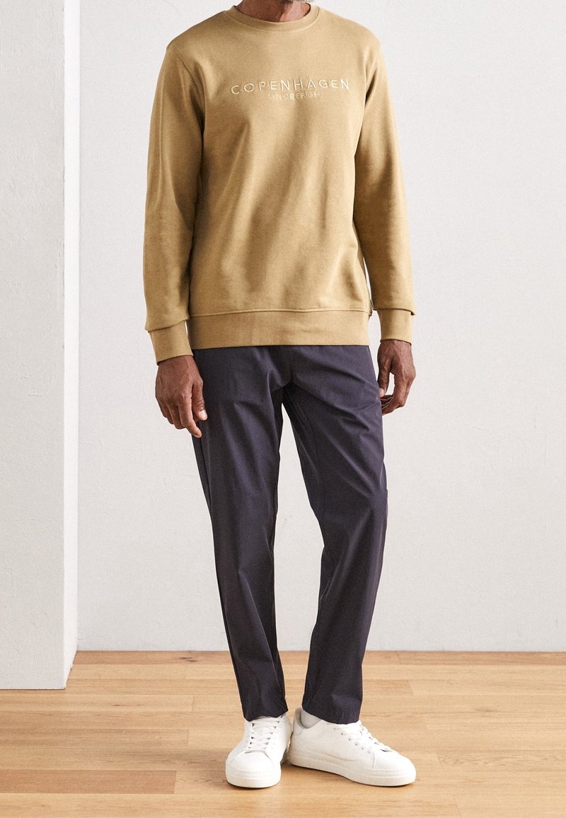 Sweat-shirt marron avec le texte "Copenhagen", associé à un pantalon foncé effilé et des baskets blanches. Tissu lisse, design simple, coupe décontractée.