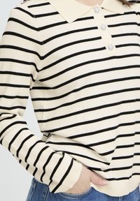 Femme portant une chemise beige à manches longues avec des rayures noires horizontales et des boutons décoratifs, main dans la poche d'un jean bleu.