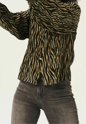 Personne portant une chemise à manches longues rayée tigre et un jean noir délavé, levant les bras, avec un fond clair.