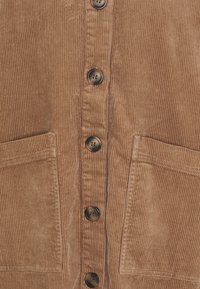 Chemise en velours côtelé marron avec des rayures verticales, comportant cinq boutons foncés et deux poches avant. Le tissu a une apparence texturée.