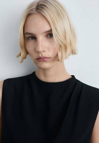 Gros plan d'une personne avec des cheveux blonds courts et ondulés, portant un haut sans manches noir. L'arrière-plan est neutre et doucement éclairé.