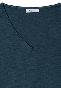 Pull col en V bleu sarcelle foncé avec texture tricotée et étiquette cousue portant la mention "Cecil" à l’intérieur du col.