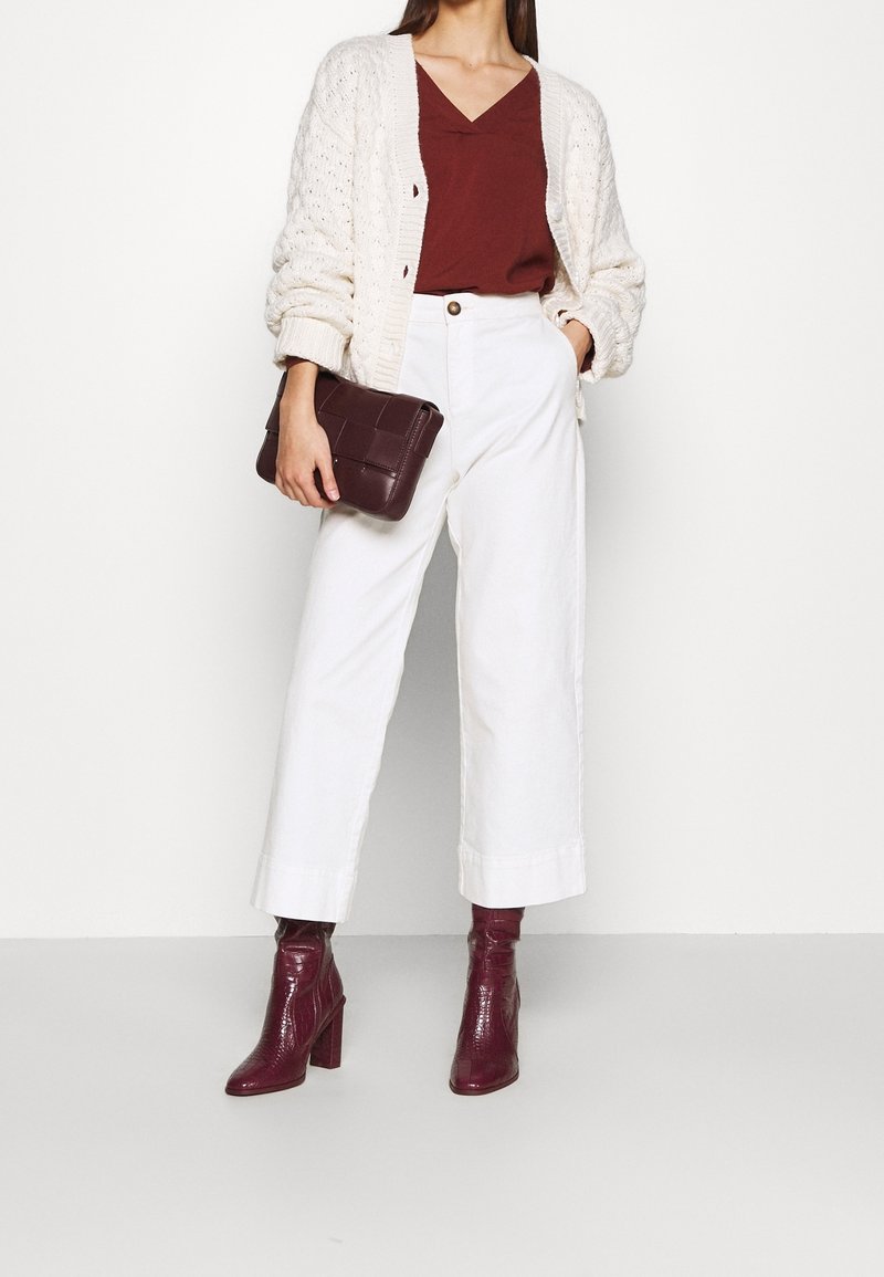 Femme portant un cardigan en maille crème par-dessus un haut bordeaux, un pantalon blanc à larges jambes, des bottines bordeaux à talons et tenant une pochette bordeaux.