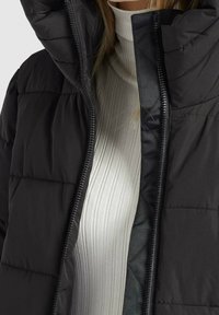 Persona vestida con una chaqueta acolchada negra con la cremallera abierta sobre un jersey de cuello alto blanco con canalé.