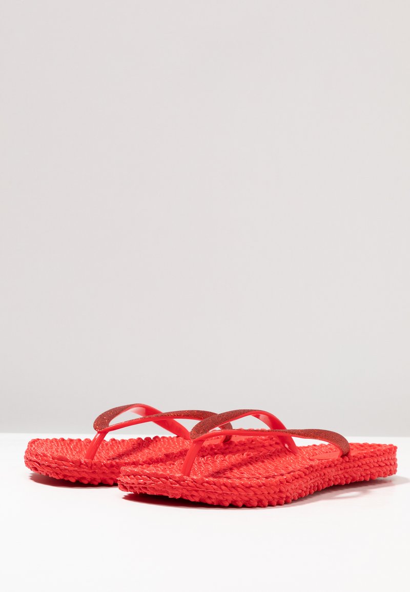 Een paar rode gevlochten slippers met gestructureerde zolen en dunne glinsterende bandjes op een witte ondergrond tegen een neutrale achtergrond.