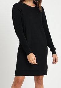 Svart långärmad klänning med ribbat mönster på ärmarna, rund halsringning och en solid kropp. Skräddarsydd passform med stickad kvalitet.