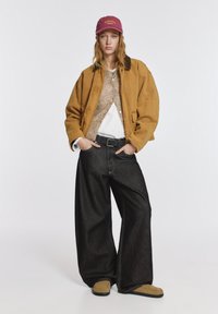 Barna túlméretezett dzseki, bézs pulóver, fehér ing, fekete bőszárú farmernadrág és barna bebújós cipők, kiegészítve egy mályvaszínű sapkával "INDIANA" felirattal.