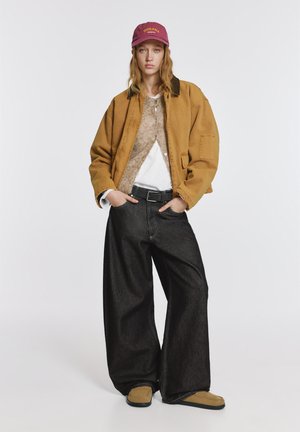 Bruine oversized jas, beige trui, wit overhemd, zwarte broek met wijde pijpen en bruine instapschoenen, aangevuld met een bordeauxrode pet met de tekst "INDIANA".