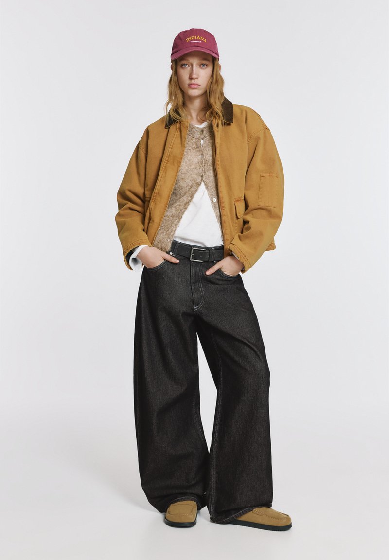 Barna túlméretezett dzseki, bézs pulóver, fehér ing, fekete bőszárú farmernadrág és barna bebújós cipők, kiegészítve egy mályvaszínű sapkával "INDIANA" felirattal.