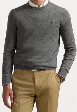 Mężczyzna ubrany w szary sweter z okrągłym dekoltem na białą koszulę z kołnierzykiem, beżowe spodnie i zegarek na nadgarstku, z jedną ręką w kieszeni, na jednolitym tle.