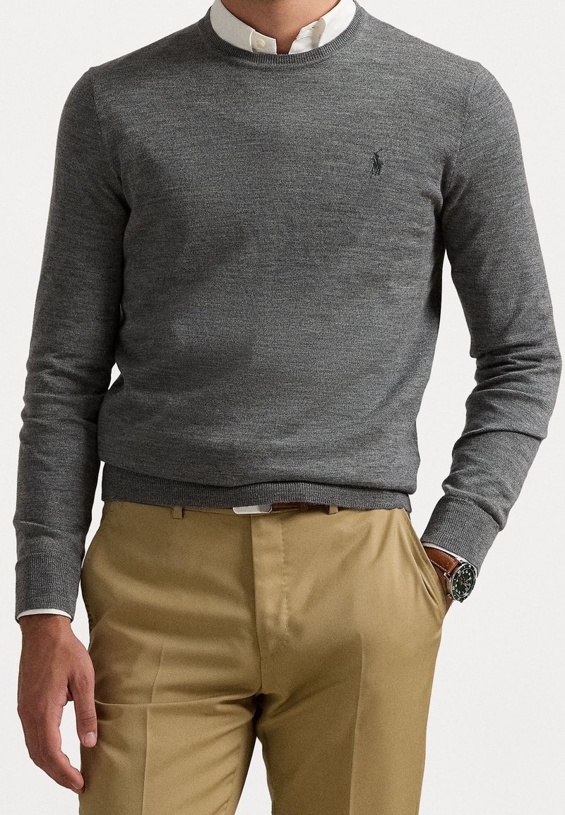 Homme portant un pull ras du cou gris sur une chemise blanche à col, avec un pantalon beige et une montre-bracelet, une main dans la poche, sur un fond uni.