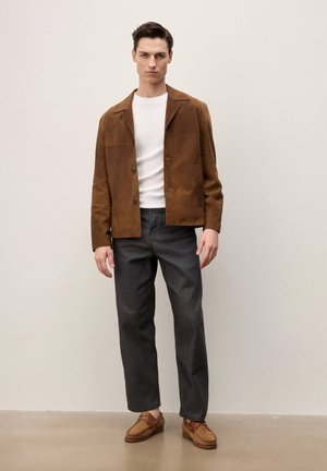 Jeune homme debout portant une veste en daim marron, un t-shirt blanc, un pantalon noir et des mocassins en daim marron, sur un fond uni.