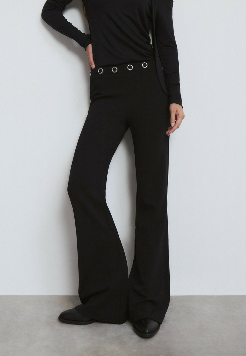 Pantalons évasés noirs avec une taille haute ornée d'accents en cercle argentés. Fabriqués en tissu lisse et extensible. Associés à un haut noir à manches longues.