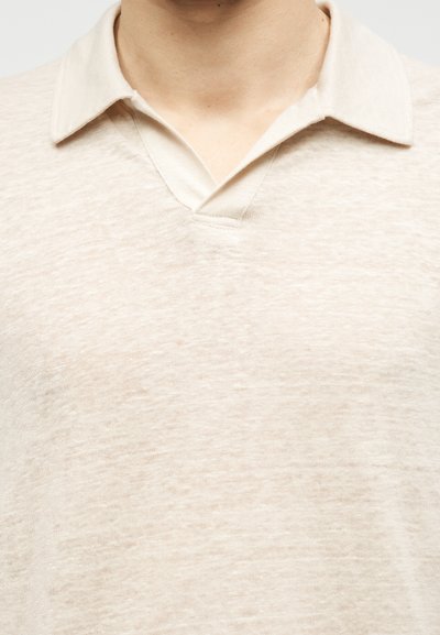 Gros plan d'une personne portant un polo beige clair texturé, col ouvert et sans boutons visibles.