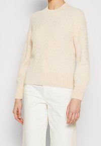 Kvinna som bär en ljusbeige strukturerad stickad tröja med lång ärm och vita byxor, stående mot en enfärgad bakgrund.