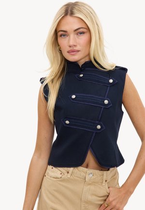 Femme blonde portant un gilet sans manches style militaire bleu marine avec des boutons argentés et un pantalon beige, debout sur fond blanc.