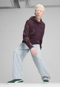 Felpa con cappuccio tie-dye color bordeaux con un piccolo logo, abbinata a pantaloni jeans larghi color blu chiaro e sneaker in suede verde con suole bianche.