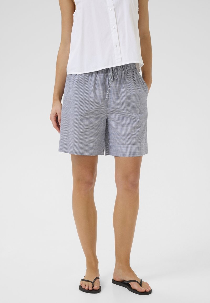 Femme portant un haut blanc sans manches boutonné, un short rayé bleu et blanc à cordon, et des tongs noires, debout les mains dans les poches.