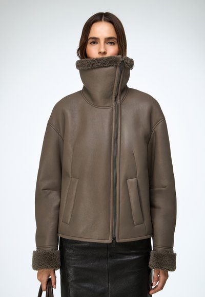 Hnedá kožená bunda s vysokým golierom, zapínaním na zips a dvoma prednými vreckami. Má kožušinou lemované manžety a hladkú povrchovú štruktúru.