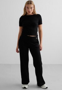 Schwarzes, kurzes T-Shirt und weit geschnittene Hose, beide aus weichem Material. Verfügt über dezente Akzente. Getragen mit weißen und schwarzen Sneakers.