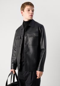 Joven con una chaqueta de cuero negra sobre un jersey negro, sosteniendo una bolsa negra, de pie contra un fondo claro y liso.
