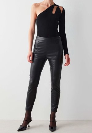 Zwarte one-shoulder bodysuit met een uitsneden detail en lange mouw; gecombineerd met nauwsluitende zwarte leren broek. Voorzien van spitse zwarte hakken.