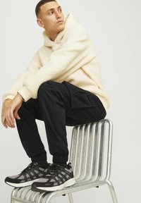 Cream fuzzy hoodie, black cargo pants with pockets, and gray/black sneakers. Sitting on a striped metal chair against a plain background.
