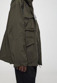 Olijfgroen, lichtgewicht jack met een capuchon, meerdere zakken, ritssluiting en elastische manchetten. Gladde textuur met een matte afwerking.