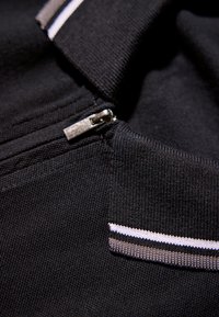 Närbild av en svart tygdragkedja med en silverfärgad dragknapp, intill en svart ribbad krage med vita och grå ränder.