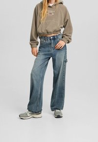 Leikattu oliivinvihreä huppari, jossa kiristysnauha, yhdistetty leveälahkeisiin sinisiin farkkuihin. Harmaat ja mustat tennarit, joissa kuvioidut pohjat. Yksinkertainen, rento tyyli.