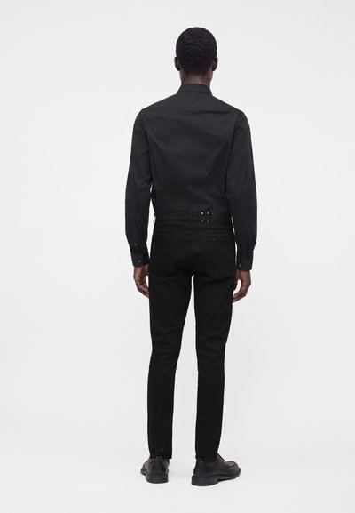 Uomo in piedi di spalle, indossa una camicia nera a maniche lunghe, pantaloni neri con cintura e scarpe eleganti nere su uno sfondo semplice.