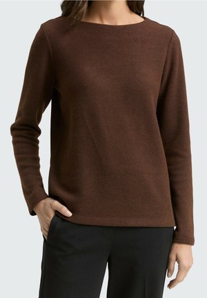 Mujer con un top de punto marrón de manga larga y cuello redondo y pantalones negros, con una mano en el bolsillo, sobre un fondo liso.