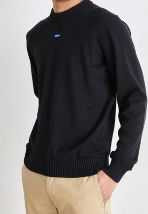 Mann trägt einen schwarzen Hugo-Pullover mit gerippten Bündchen und Saum sowie beigefarbene Hose, mit einer Hand in der Tasche, vor einem weißen Hintergrund stehend.