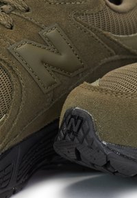 Zamszowe i siateczkowe sneakersy w oliwkowej zieleni z czarnymi, teksturowanymi podeszwami, wyposażone w duże, zszywane logo "N" z boku i na podeszwie.