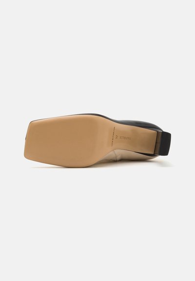 Zwarte en beige hoge hak schoen met een chunky hak, een soepele leren bovenkant en een rubberen zool, met een zwarte strap aan de achterkant.