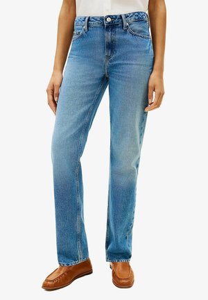 Femme portant un jean bleu taille mi-haute, coupe droite, et des mocassins en cuir marron, debout sur fond blanc.