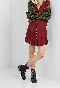 Burgundininen mekko, jossa on V-kaula-aukko ja napitusyksityiskohtia, yhdistetty vihreään camo-takkiin; mustat nauhakenkäankkal bootsit, joissa on piikikkäitä koristeita.