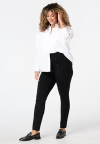 Vrouw met krullend haar, gekleed in een witte overhemd met knopen, zwarte knopenbroeken en zwarte studsloafers, staande en lachend met haar hand die haar aanraakt.