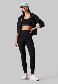 Zwarte sportbeha met logo, zwarte high-waisted leggings en een zwarte hoodie met ritssluiting. Een lichtblauwe pet en witte sneakers maken de look af.