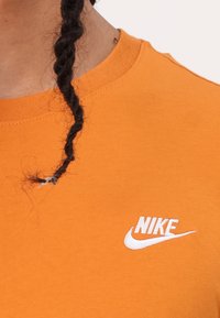 Oranžové bavlněné tričko s kulatým výstřihem, na kterém se nachází bílé logo Nike na levé straně hrudi. Tkanina má hladkou texturu.