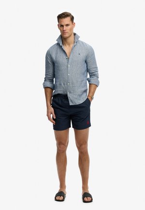 Mann trägt hellblaues Hemd mit aufgeklapptem Kragen, marineblaue Shorts, schwarze Sandalen, steht mit einer Hand in der Tasche.