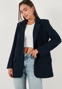 Tengerész kék blézer, amely testhezálló szabású, strukturált hajtókákkal és két elülső zsebbel rendelkezik. Egy bordázott fehér crop top fölött viselve, világos farmernadrággal párosítva.