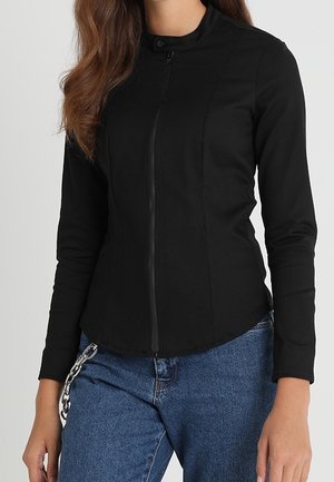 Vrouw draagt een nauwsluitend zwart langarmshirt met ritssluiting en een hoge kraag, gecombineerd met een blauwe spijkerbroek en een ketting die uit de zak hangt.