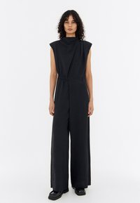 Black sleeveless jumpsuit with an asymmetric design, elastic waist, and wide legs. Made from textured fabric with subtle speckles.