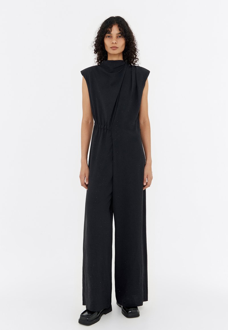 Black sleeveless jumpsuit with an asymmetric design, elastic waist, and wide legs. Made from textured fabric with subtle speckles.