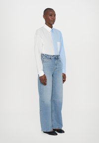 Cotton two-tone shirt featuring a white and light blue design, with a front pocket; paired with wide-leg light blue jeans and black shoes.