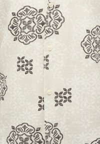 Ανοιχτό μπεζ ύφασμα με μαύρα, περίτεχνα συμμετρικά λουλουδάτα και γεωμετρικά σχέδια, και τρία ορατά κρεμ κουμπιά τοποθετημένα κάθετα.