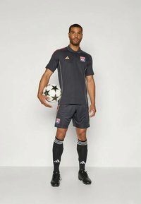 Σκούρο γκρίζο ποδοσφαιρικό σετ με κοντά μανίκια, που περιλαμβάνει γιακά, κόκκινες και χρυσές λεπτομέρειες και λογότυπο. Μαύρα σορτς και κάλτσες με τρεις λευκές ρίγες. Κρατώντας μια ασπρόμαυρη μπάλα ποδοσφαίρου.