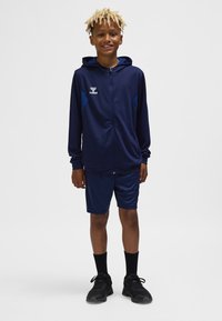 Marineblå atletisk træningsdragt med lynlåshætte, kontrasterende blå accenter og matchende shorts. Bæres med sorte sneakers og sokker.