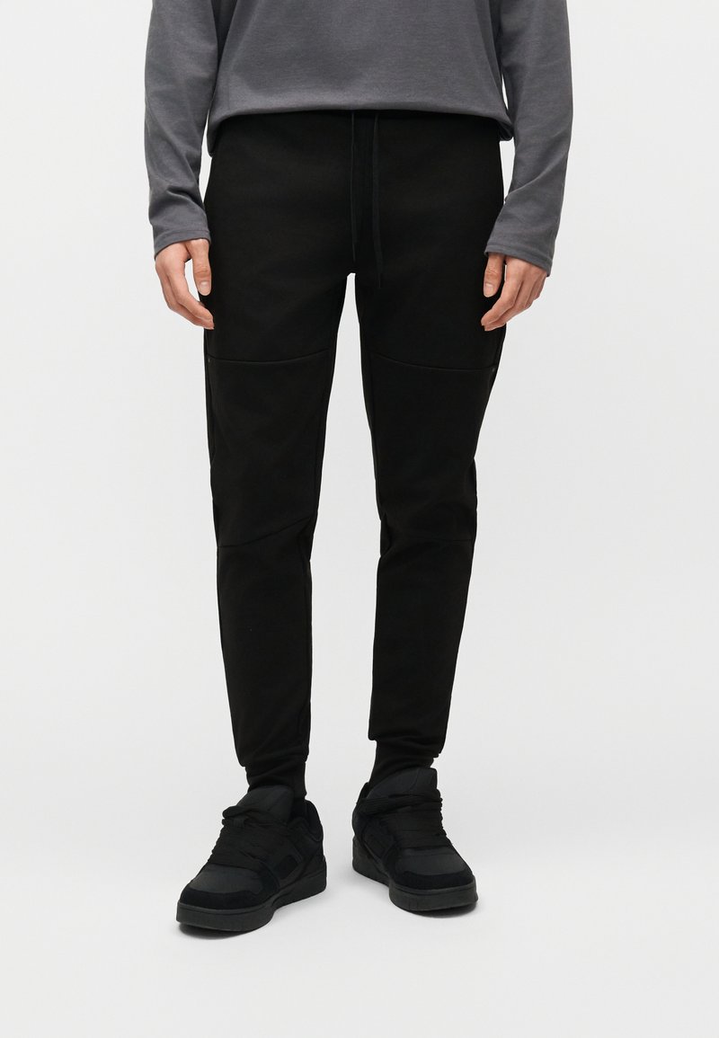 Άνδρας που φοράει μαύρο στενό παντελόνι jogger με κορδόνι στη μέση και μαύρα αθλητικά παπούτσια, στέκεται μπροστά σε απλό φωτεινό φόντο.